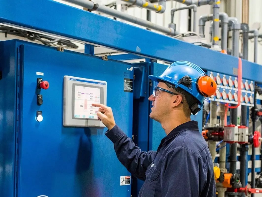 Travailleur portant un casque de sécurité et manipulant un écran tactile sur un équipement industriel.