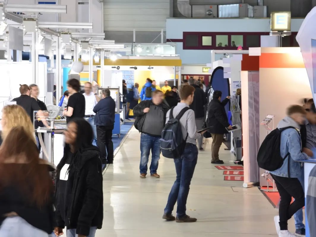 Visitors at a busy indoor trade show with exhibition booths and walking attendees.